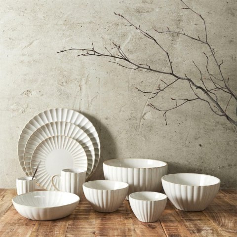 Zestaw beżowych naczyń ceramicznych z serii Beju, w tym miski, talerze i kubek z uchem pod betonową ścianą.